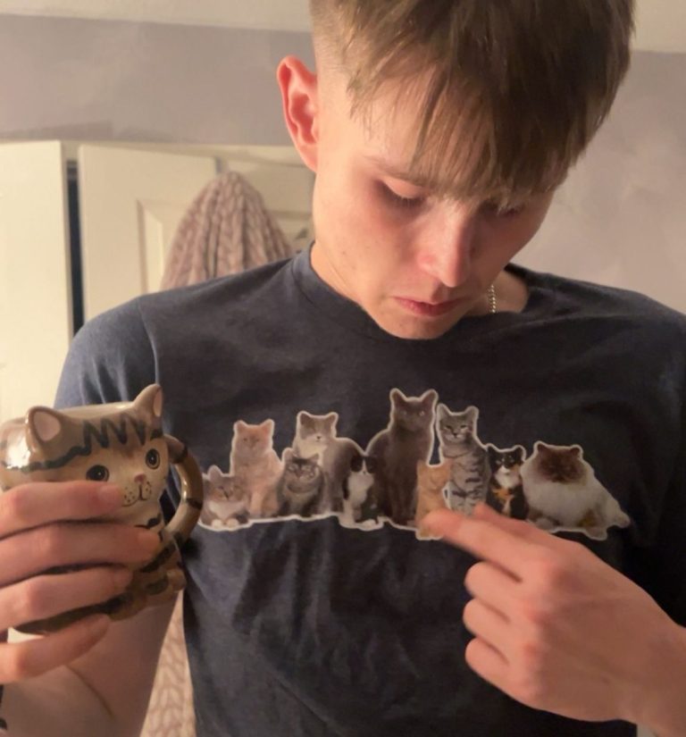 A person points to a cat-themed shirt while holding a cat-shaped mug.